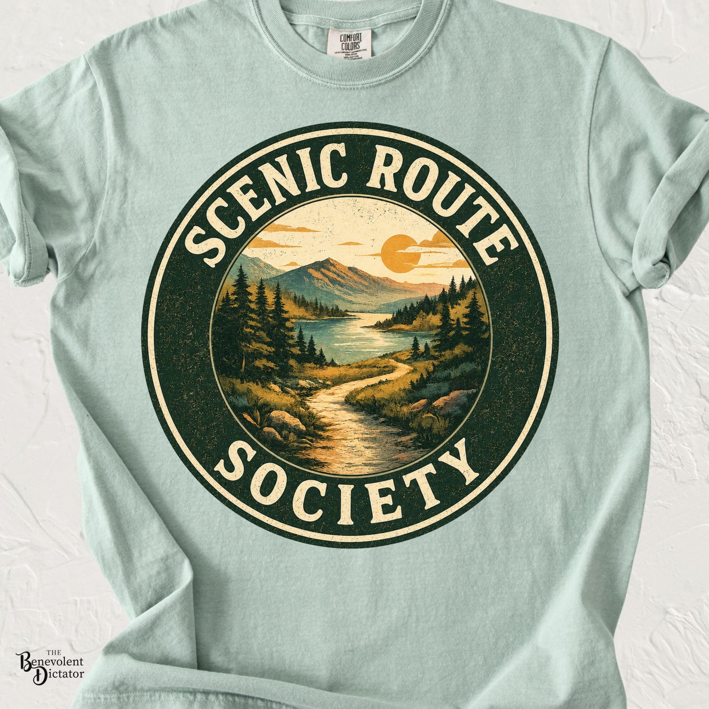 Vintage-style circular landscape featuring a winding trail, mountains, lake, and the words "Scenic Route Society" in distressed retro typography on a transparent background.