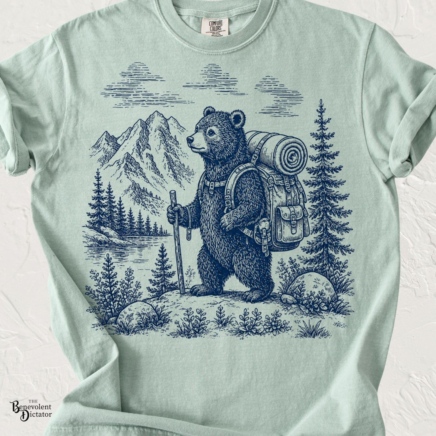 Vintage blue line-art illustration of a backpacking bear standing beside a mountain lake with pine trees on a Comfort Colors shirt.