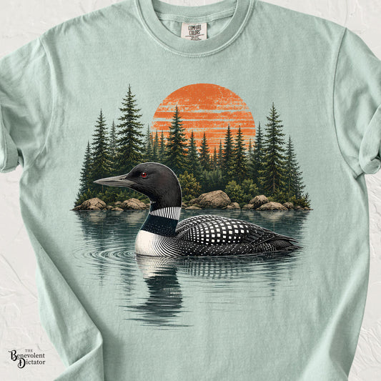 Vintage common loon graphic t-shirt featuring a loon swimming on a peaceful lake surrounded by evergreen forest and a distressed orange sunset.