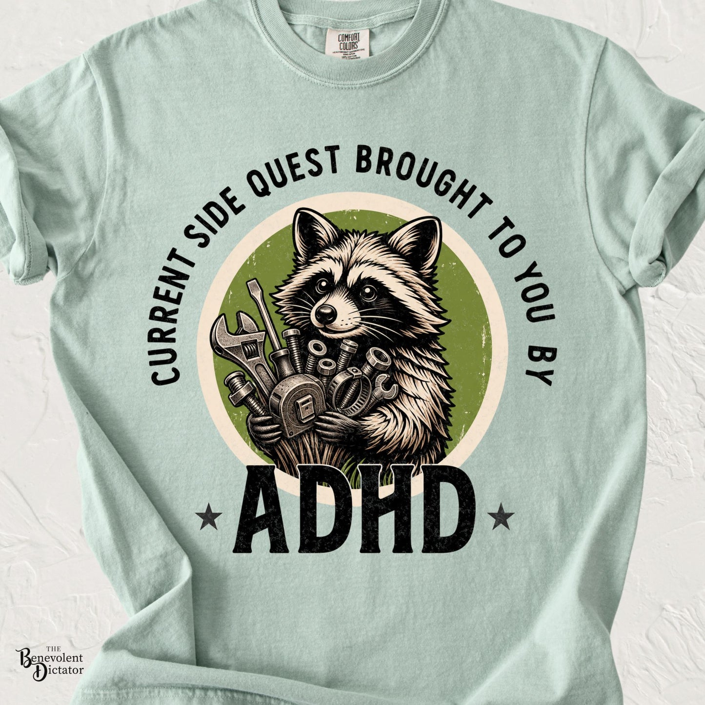 Comfort Colors Bay green t-shirt featuring a proud raccoon holding a collection of tools and hardware with the phrase "Current Side Quest Brought to You by ADHD" in bold vintage lettering.