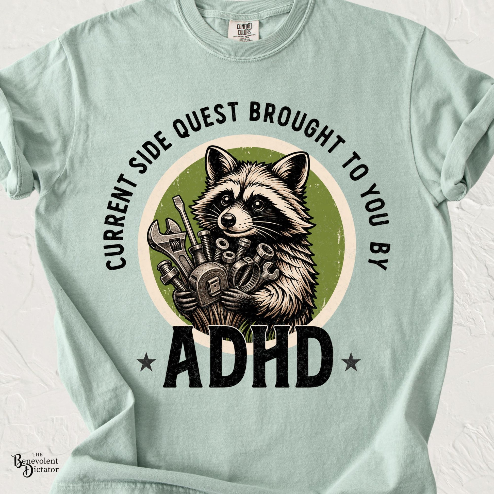 Comfort Colors Bay green t-shirt featuring a proud raccoon holding a collection of tools and hardware with the phrase "Current Side Quest Brought to You by ADHD" in bold vintage lettering.