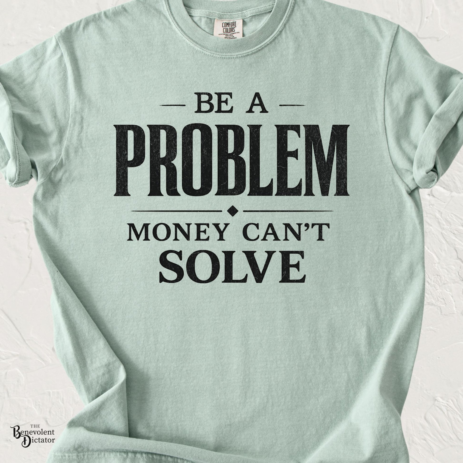 Mint green graphic t-shirt with bold typography reading “Be a Problem Money Can’t Solve” in distressed black lettering.