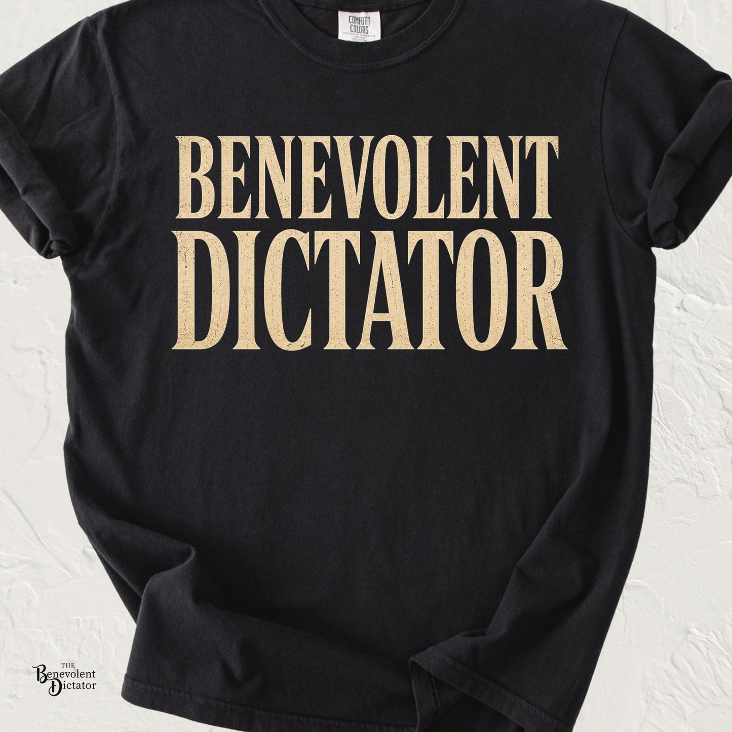 graphic t-shirt featuring bold vintage serif typography reading Benevolent Dictator in distressed cream lettering.