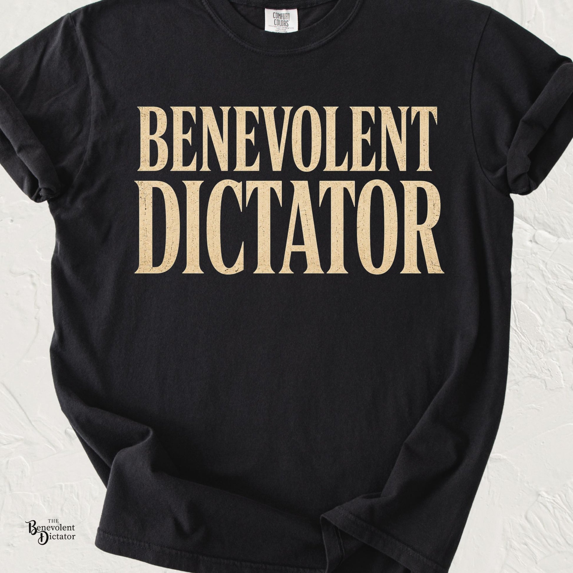 graphic t-shirt featuring bold vintage serif typography reading Benevolent Dictator in distressed cream lettering.