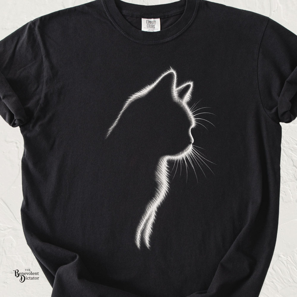 Black Comfort Colors t-shirt featuring a minimalist white illuminated outline of a cat in profile, with delicate glowing fur and whisker details against the dark fabric.