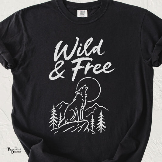 Black Comfort Colors t-shirt featuring a distressed white “Wild & Free” design with a howling wolf, moon, mountains, and pine trees.