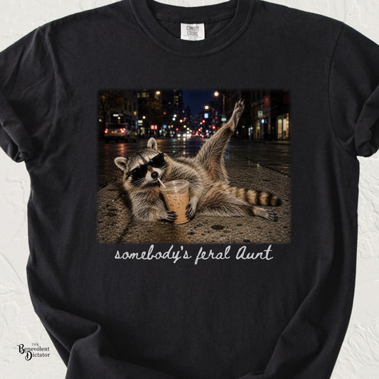 Black Comfort Colors t-shirt featuring a sunglasses-wearing raccoon sprawled on a city street at night drinking iced coffee, with handwritten text reading “somebody’s feral Aunt.”