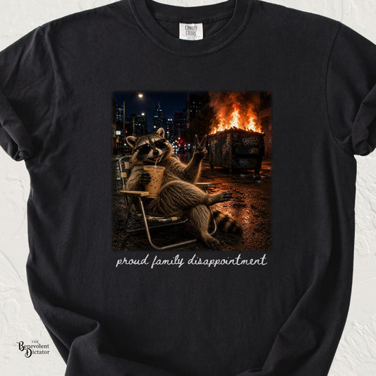 Black Comfort Colors t-shirt featuring a sunglasses-wearing raccoon lounging in a chair with iced coffee beside a burning dumpster, with text reading “proud family disappointment.”