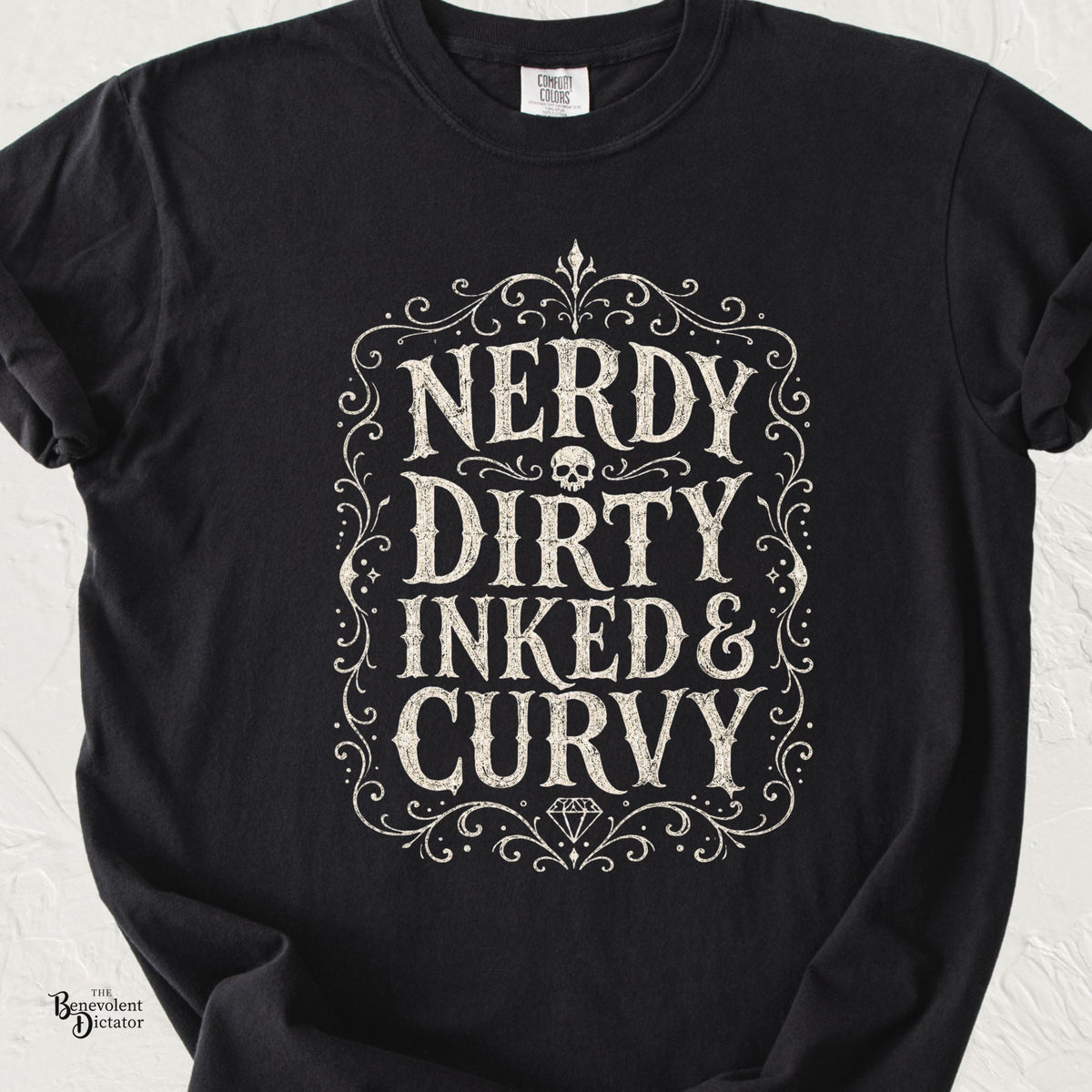 Black Comfort Colors t-shirt with distressed ivory typography reading Nerdy Dirty Inked & Curvy, surrounded by ornate vintage flourishes, a skull, and a diamond.