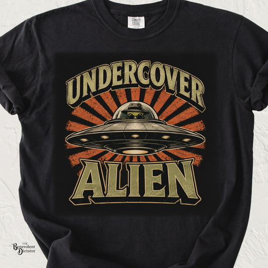 Black Comfort Colors t-shirt featuring a vintage-style UFO with a sunglasses-wearing alien, orange sunburst, and distressed typography reading Undercover Alien.