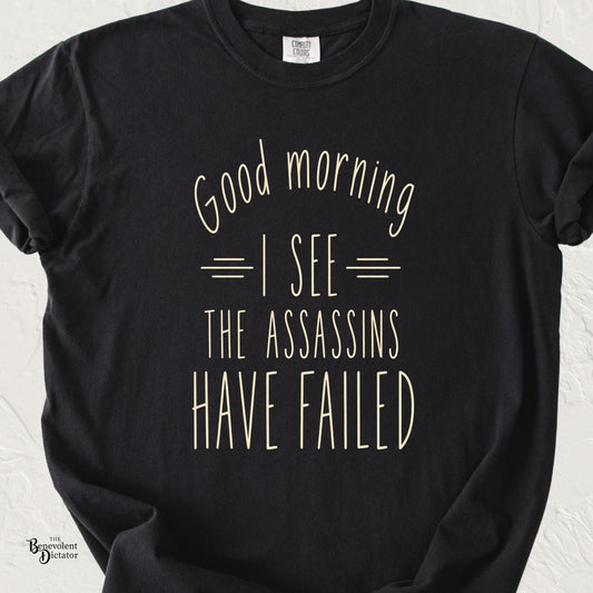 Black Comfort Colors t-shirt with minimalist cream typography reading Good Morning I See the Assassins Have Failed.