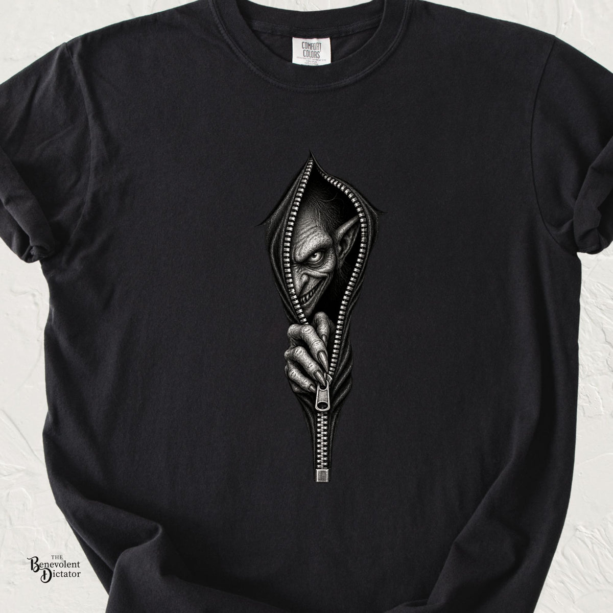 Black Comfort Colors t-shirt featuring a sinister goblin emerging through an open zipper, gripping the zipper pull from inside.