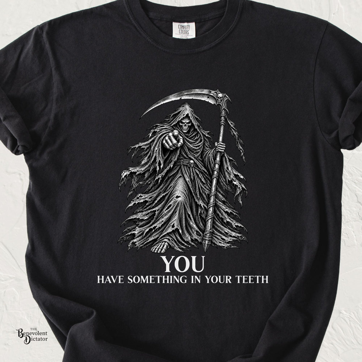 Black Comfort Colors t-shirt featuring a dark illustrated Grim Reaper pointing forward while holding a scythe above the words “You Have Something in Your Teeth.”