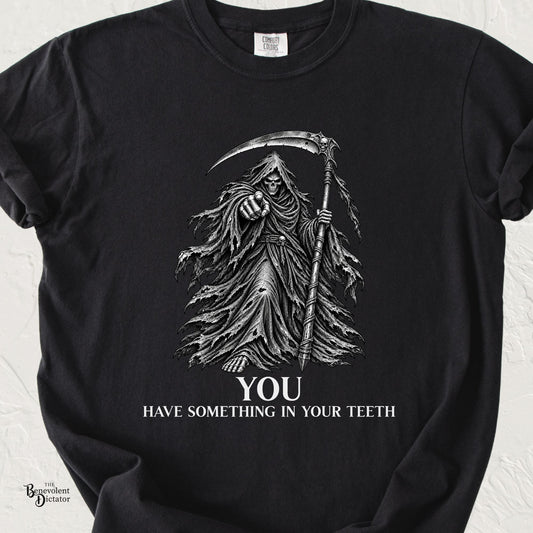 Black Comfort Colors t-shirt featuring a dark illustrated Grim Reaper pointing forward while holding a scythe above the words “You Have Something in Your Teeth.”