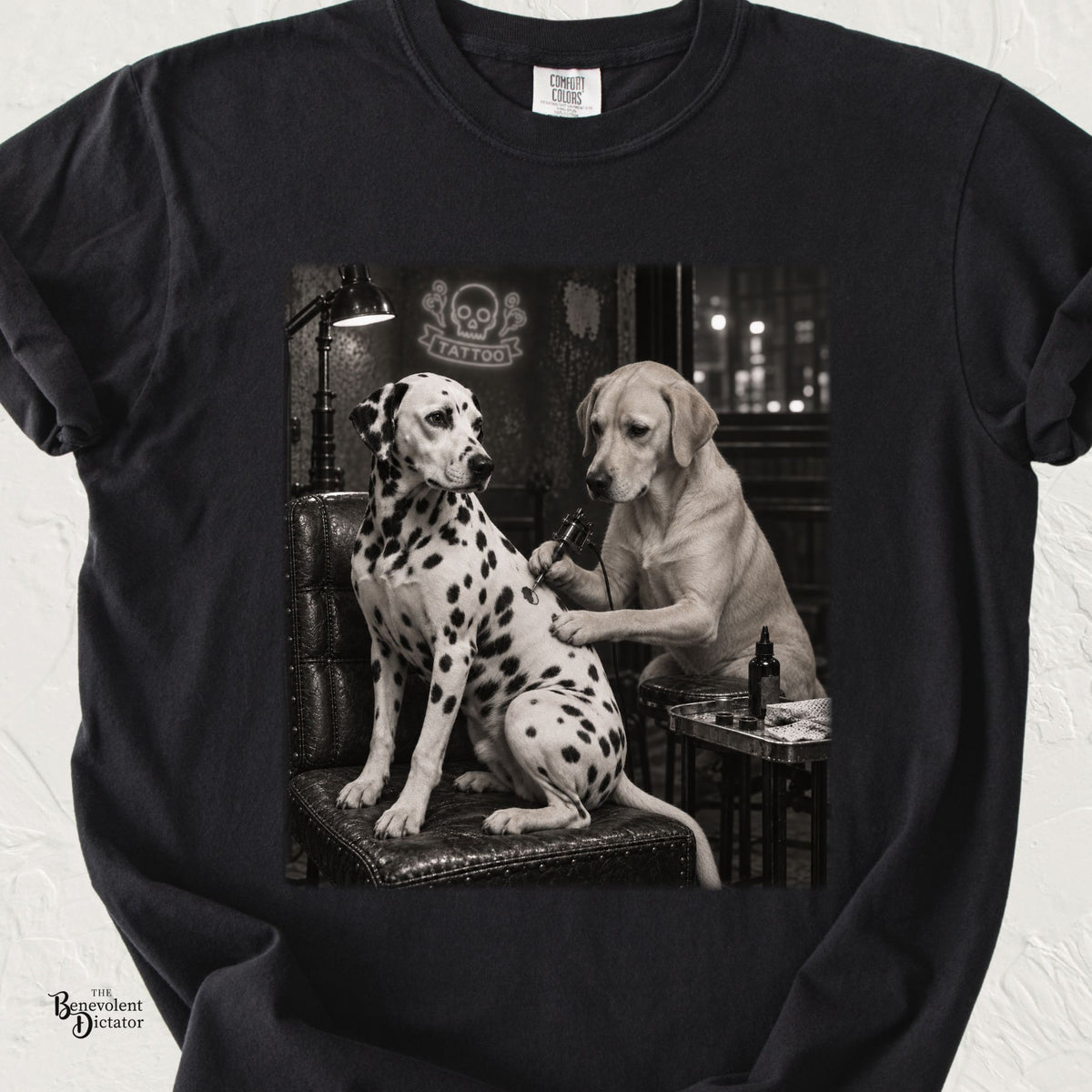 Black Comfort Colors t-shirt featuring a black-and-white scene of a Labrador tattoo artist giving a Dalmatian new spots inside a gritty nighttime tattoo parlor.