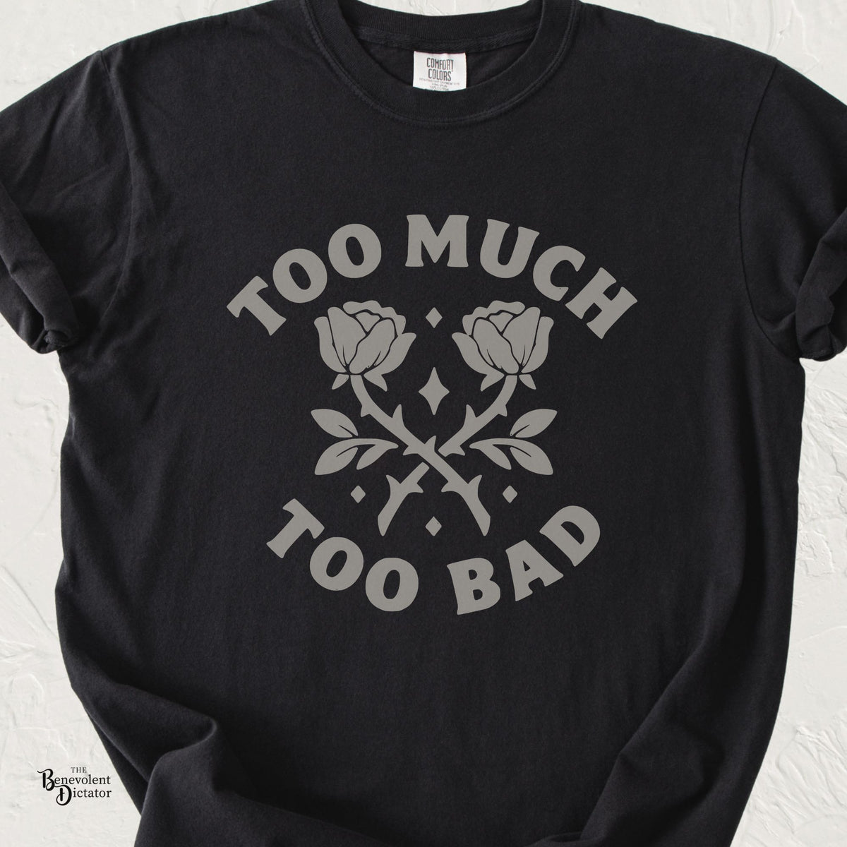 Black Comfort Colors t-shirt featuring crossed roses and small diamond accents with the phrase “Too Much Too Bad” in bold vintage lettering.