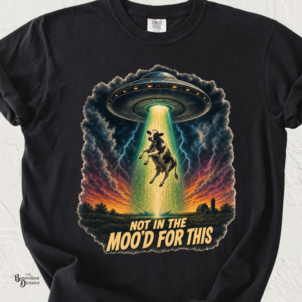Black Comfort Colors t-shirt featuring a cow being abducted by a UFO in a dramatic lightning-filled sky with the phrase “Not in the Moo’d for This.”