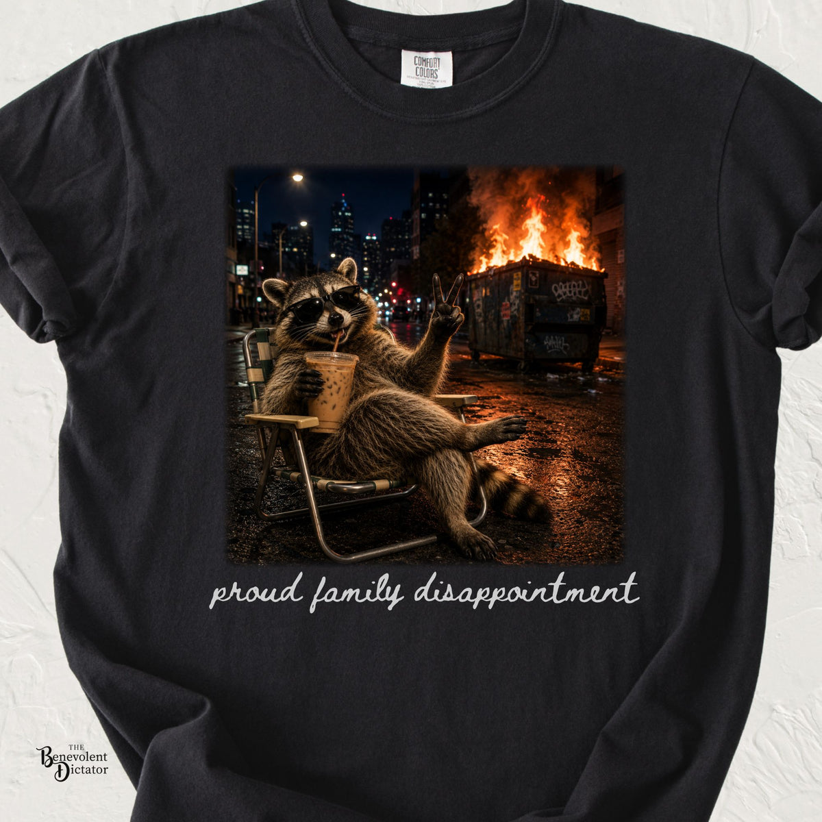 A black short-sleeve t-shirt with a graphic print of a raccoon wearing sunglasses and sitting in a chair next to a burning dumpster, with the text 'proud family disappointment' printed below.