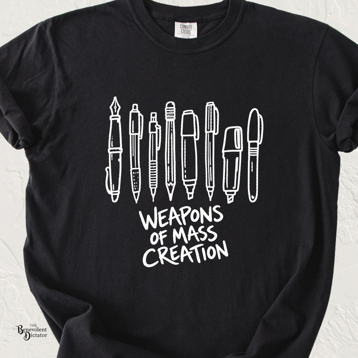 Black Comfort Colors t-shirt featuring hand-drawn pens, pencils and markers lined up above the words “Weapons of Mass Creation.”