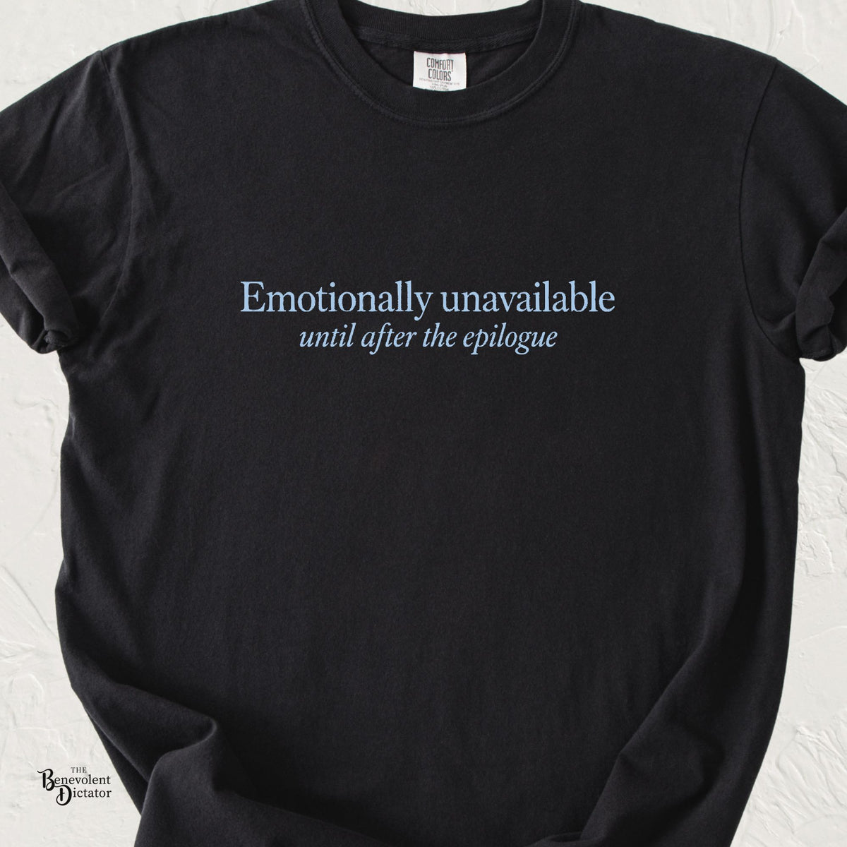 Black Comfort Colors t-shirt featuring understated baby blue serif typography reading “Emotionally unavailable” with “until after the epilogue” in smaller italic lettering beneath.