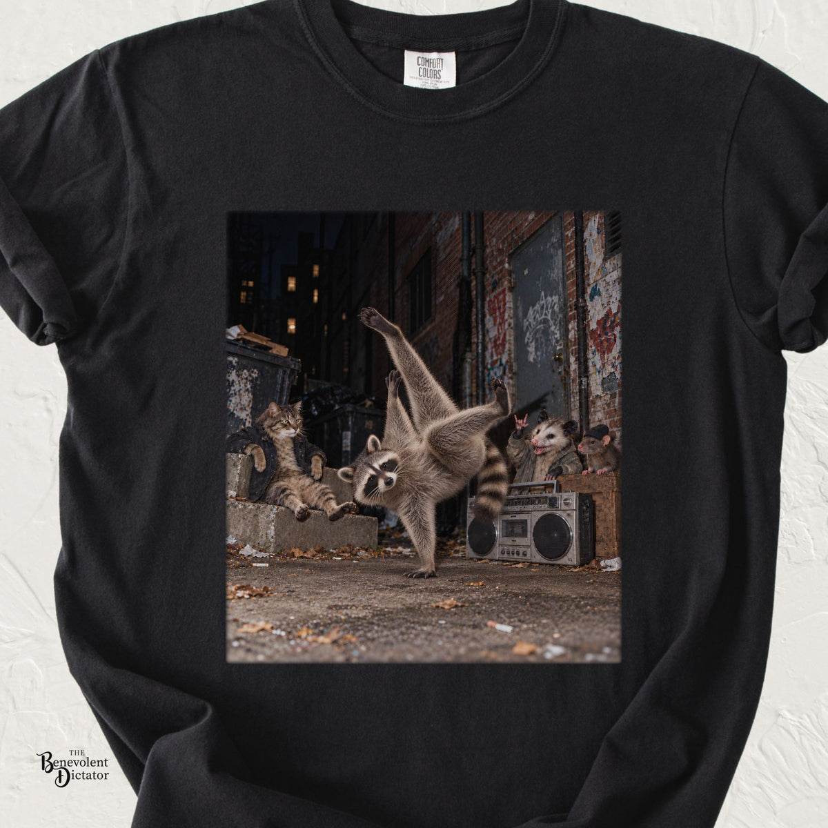 Black Comfort Colors t-shirt featuring a photorealistic nighttime urban alley scene with a raccoon breakdancing on one paw, a lounging tabby cat, and an opossum and rat cheering beside a vintage boombox.