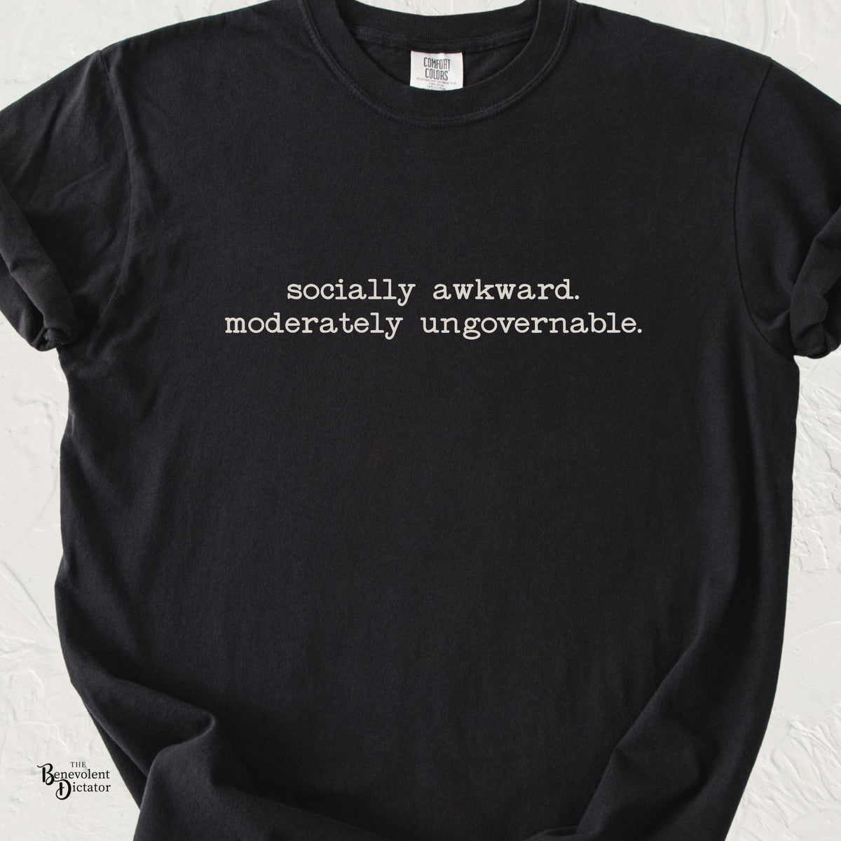Black Comfort Colors t-shirt featuring minimalist off-white typewriter-style typography reading “socially awkward. moderately ungovernable.” in two centered lines across the chest.