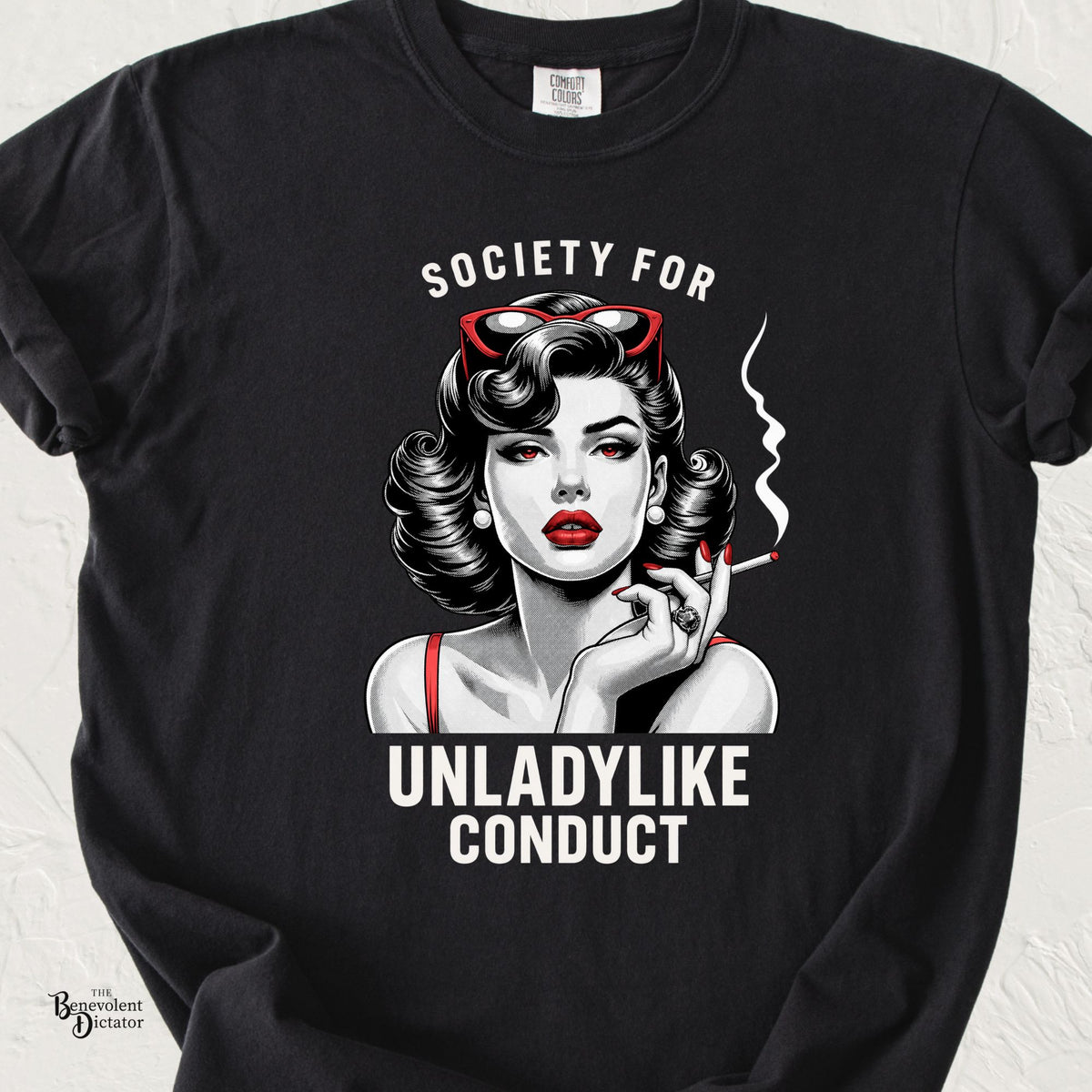 Black Comfort Colors t-shirt featuring a vintage pin-up style woman with dark curled hair, red cat-eye sunglasses, red lips and a lit cigarette. Bold white lettering reads “Society for Unladylike Conduct.”