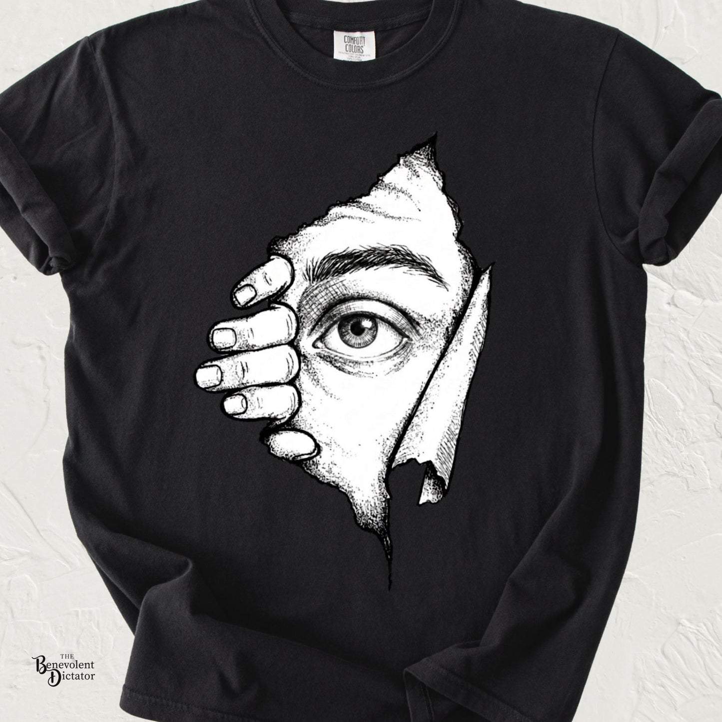 Black Comfort Colors t-shirt featuring a surreal black-and-white illustration of a hand pulling back torn paper to reveal a single curious eye peeking through.