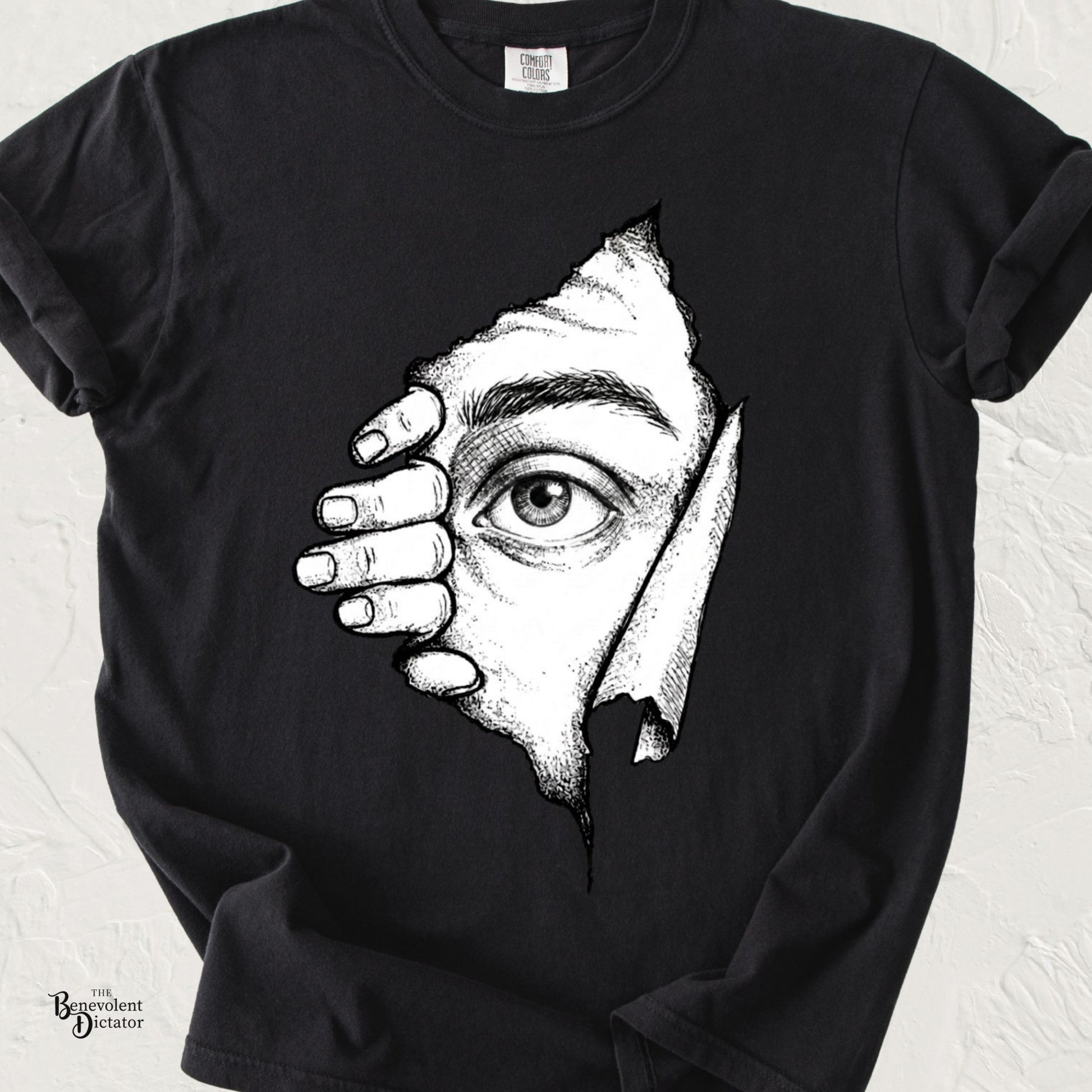 Black Comfort Colors t-shirt featuring a surreal black-and-white illustration of a hand pulling back torn paper to reveal a single curious eye peeking through.