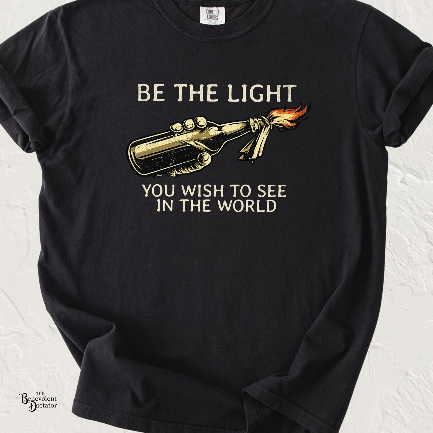 Black graphic t-shirt featuring a Molotov cocktail illustration with the phrase “Be the Light You Wish to See in the World.”