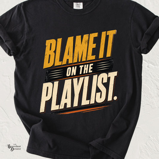 Black Comfort Colors t-shirt featuring a bold distressed typography design that reads "Blame It on the Playlist" in cream, gold, and orange vintage lettering.