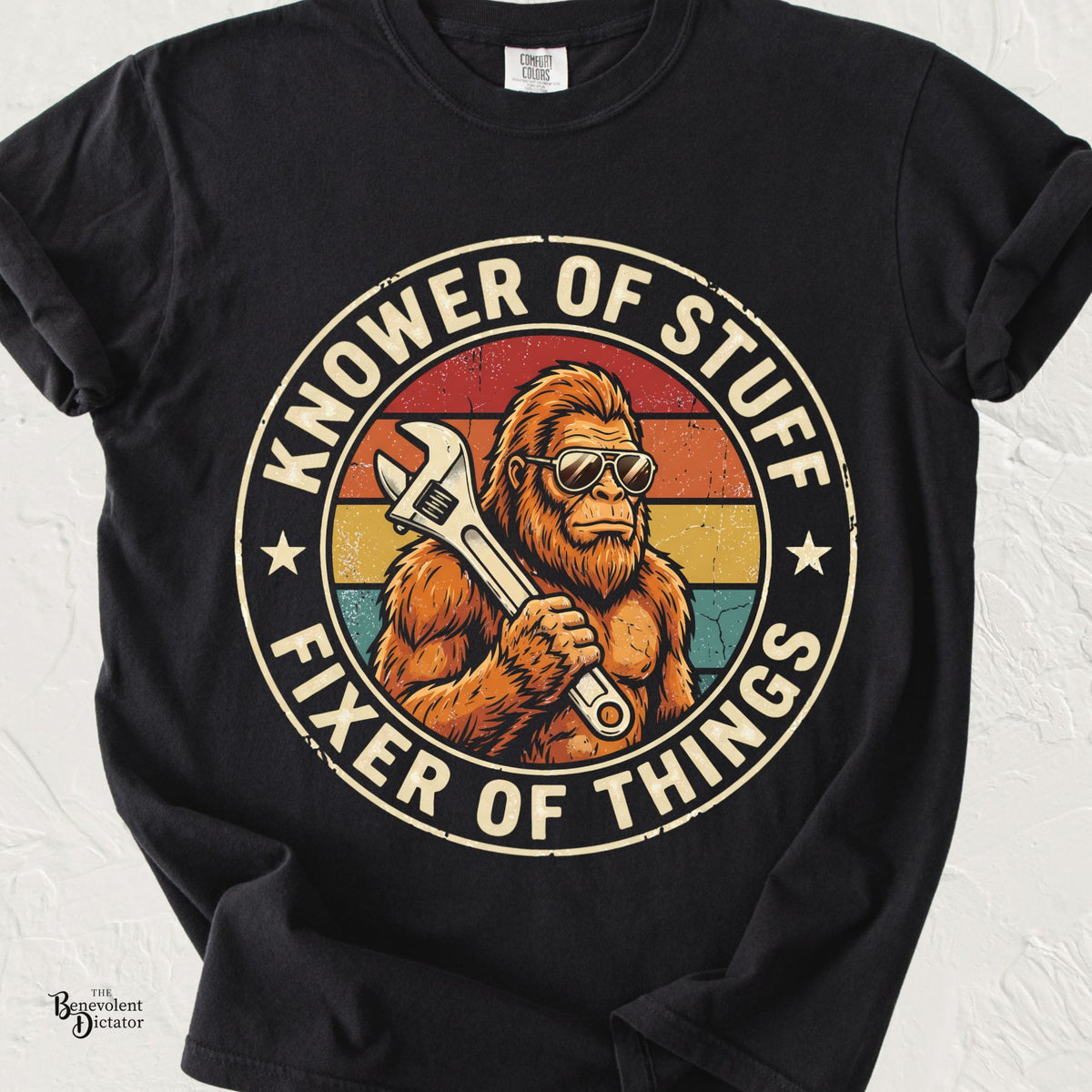 Comfort Colors black t-shirt featuring a vintage-style Bigfoot wearing aviator sunglasses and holding a wrench inside a retro sunset badge with the phrase "Knower of Stuff, Fixer of Things."