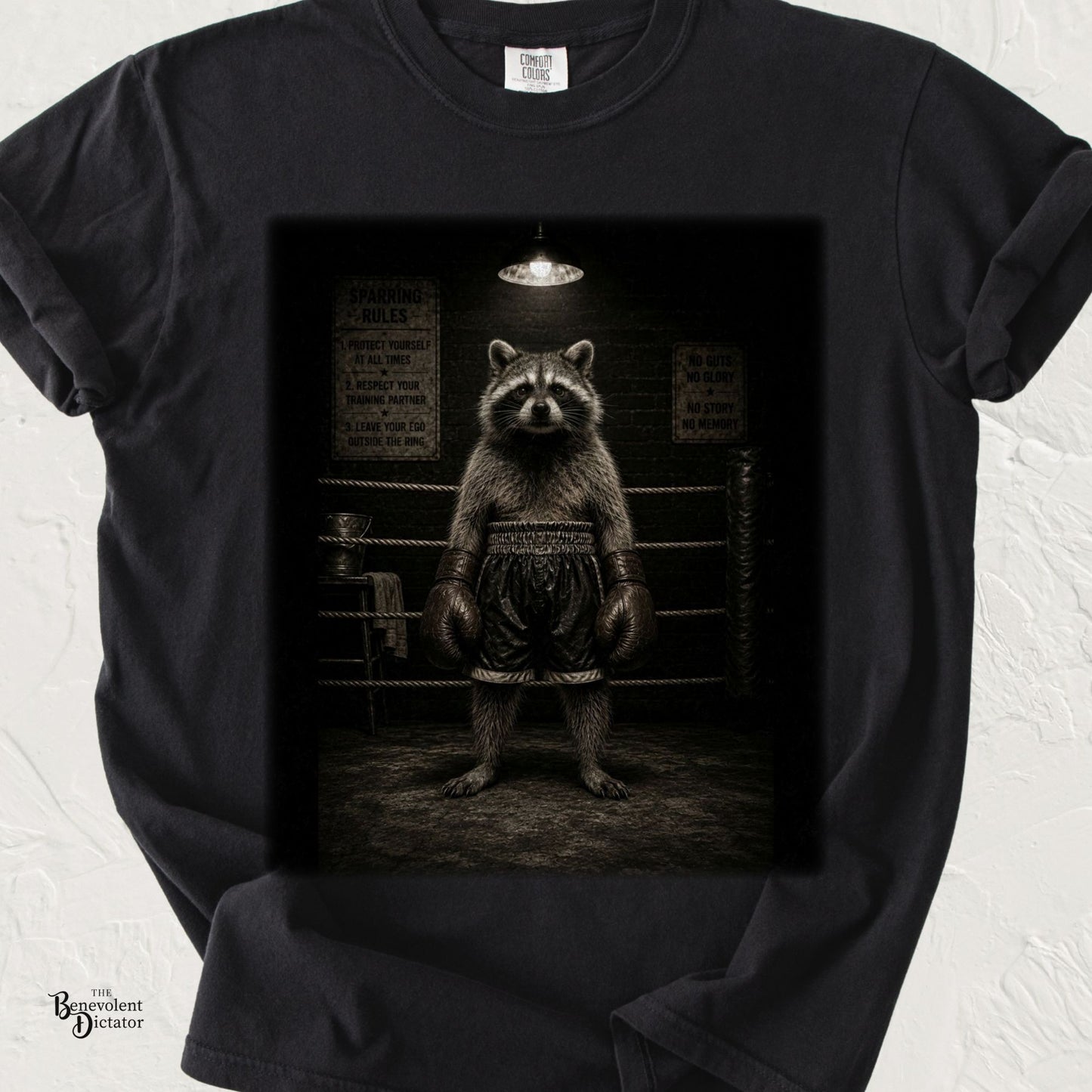 Vintage-style illustration of a determined raccoon wearing oversized boxing gloves and trunks, standing confidently in a dimly lit boxing ring on a black Comfort Colors T-shirt.