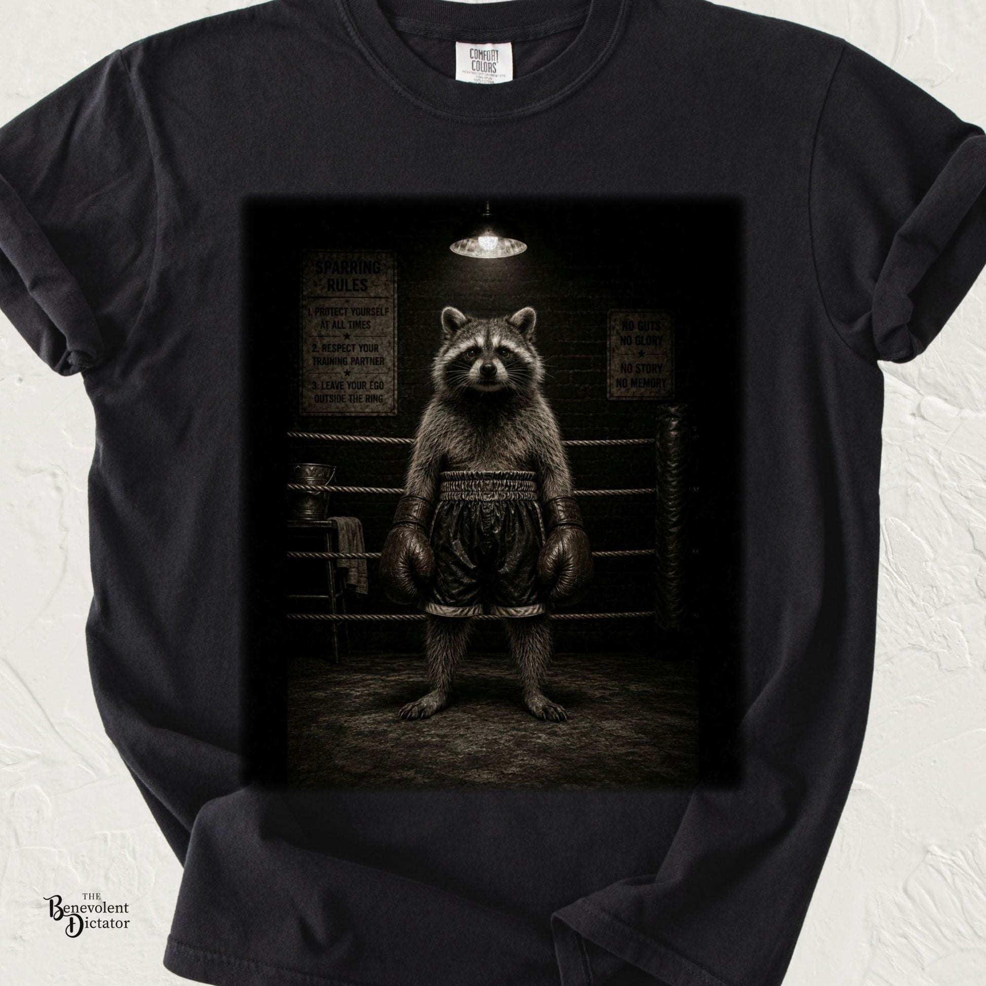 Vintage-style illustration of a determined raccoon wearing oversized boxing gloves and trunks, standing confidently in a dimly lit boxing ring on a black Comfort Colors T-shirt.