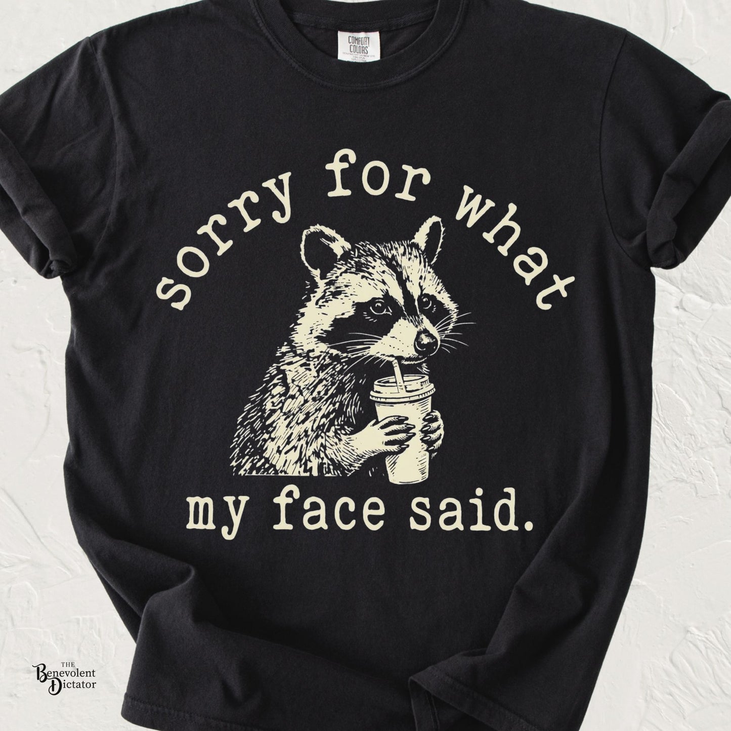 Funny raccoon T-shirt featuring a vintage-style raccoon sipping coffee with the phrase "Sorry for What My Face Said" printed in distressed cream lettering on a black Comfort Colors shirt.