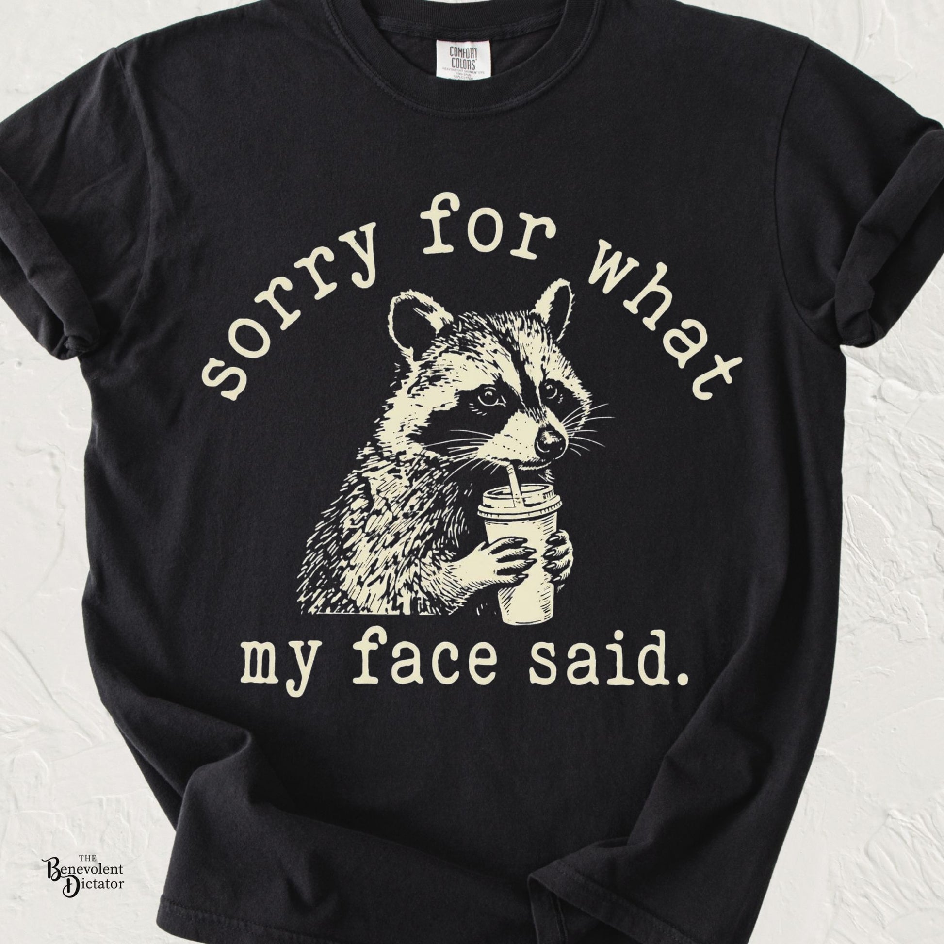 Funny raccoon T-shirt featuring a vintage-style raccoon sipping coffee with the phrase "Sorry for What My Face Said" printed in distressed cream lettering on a black Comfort Colors shirt.