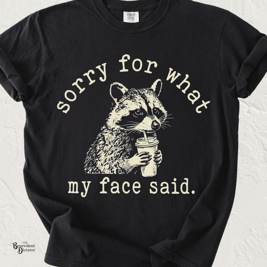 Funny raccoon T-shirt featuring a vintage-style raccoon sipping coffee with the phrase "Sorry for What My Face Said" printed in distressed cream lettering on a black Comfort Colors shirt.
