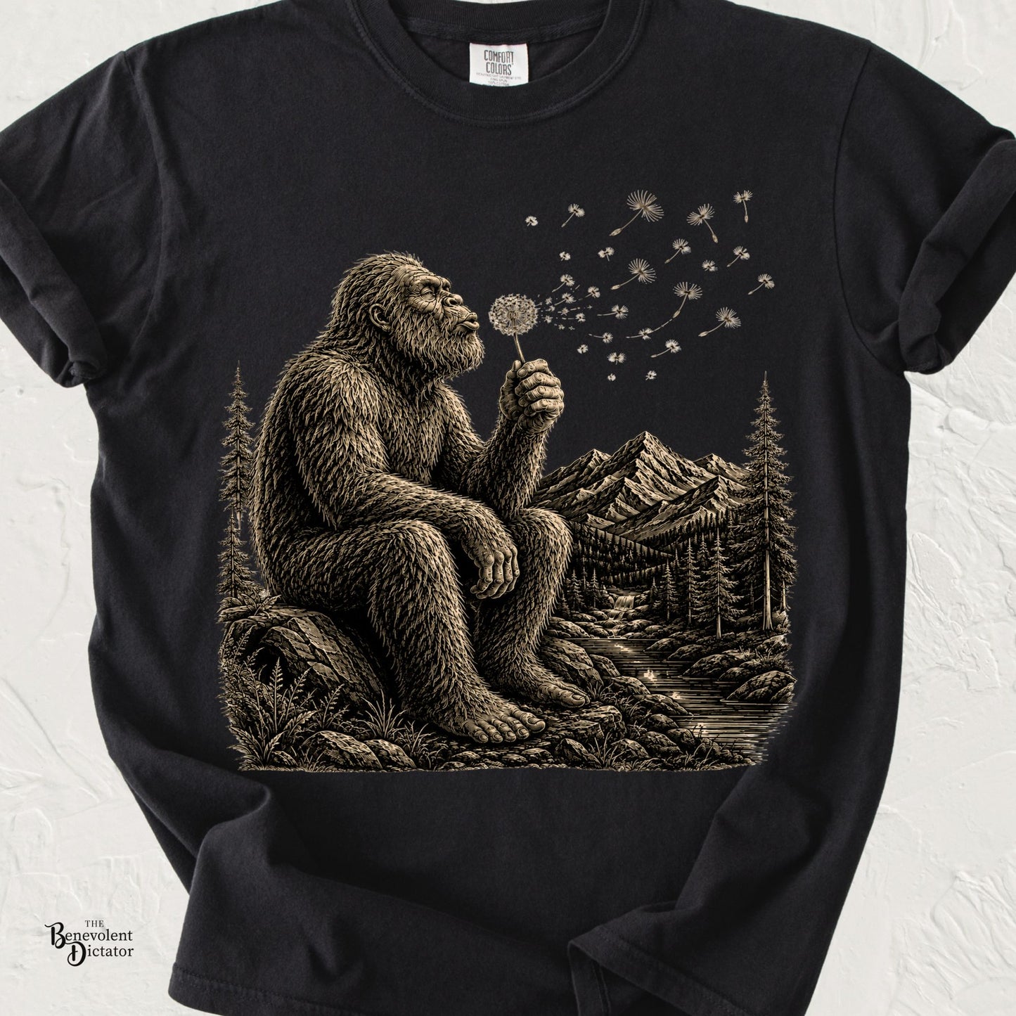 Vintage-style Bigfoot sitting on a rock blowing a dandelion with mountains, forest, and river in the background.