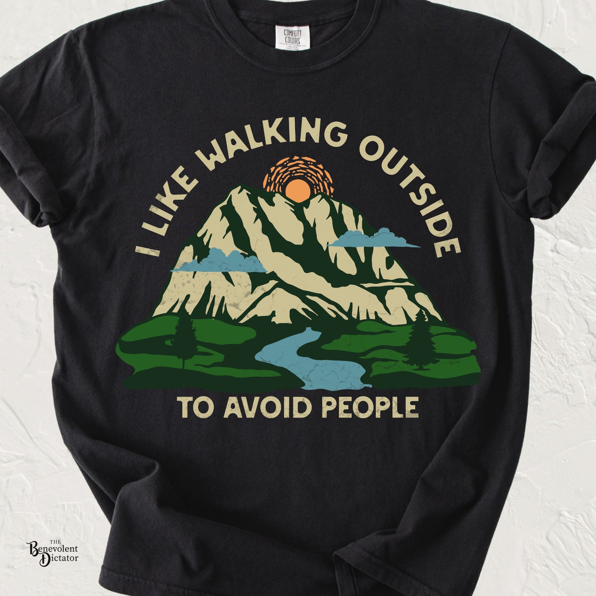 Vintage-style graphic featuring a mountain landscape with a winding river, sun, and evergreen trees with the humorous phrase "I Like Walking Outside to Avoid People."