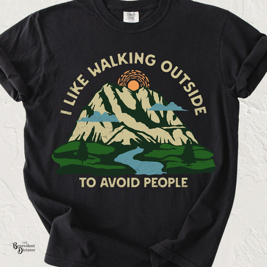 Vintage-style graphic featuring a mountain landscape with a winding river, sun, and evergreen trees with the humorous phrase "I Like Walking Outside to Avoid People."