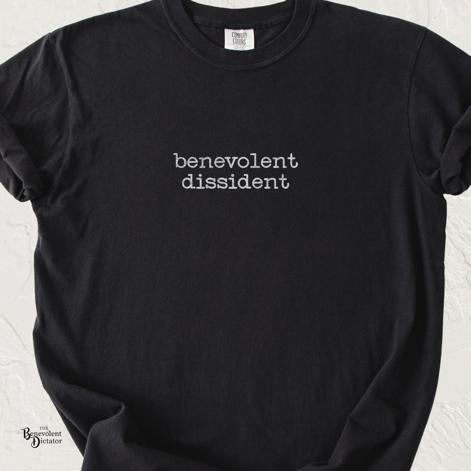 Minimalist Benevolent Dissident typography graphic in distressed lowercase typewriter lettering on a black Comfort Colors t-shirt.