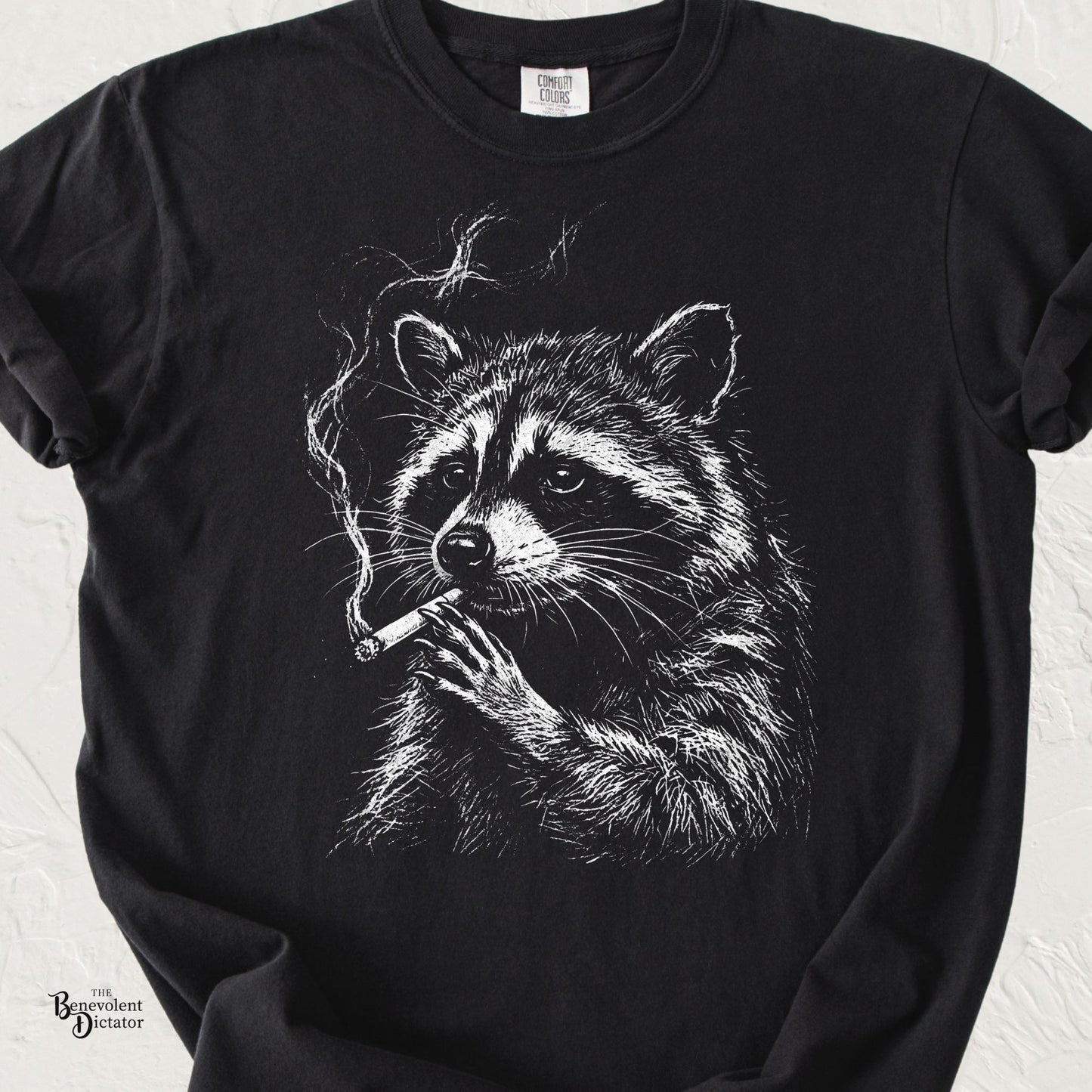 Black Comfort Colors t-shirt featuring a vintage white line-art raccoon holding a smoking cigarette with a deadpan expression.