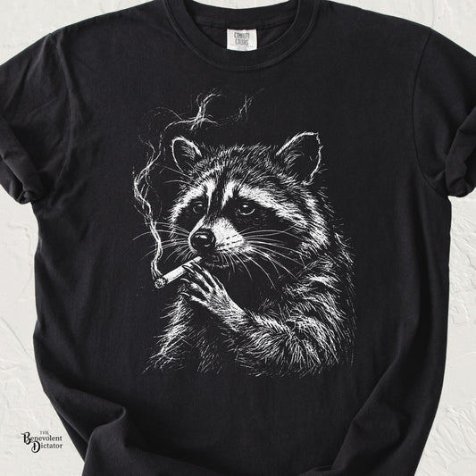 Black Comfort Colors t-shirt featuring a vintage white line-art raccoon holding a smoking cigarette with a deadpan expression.