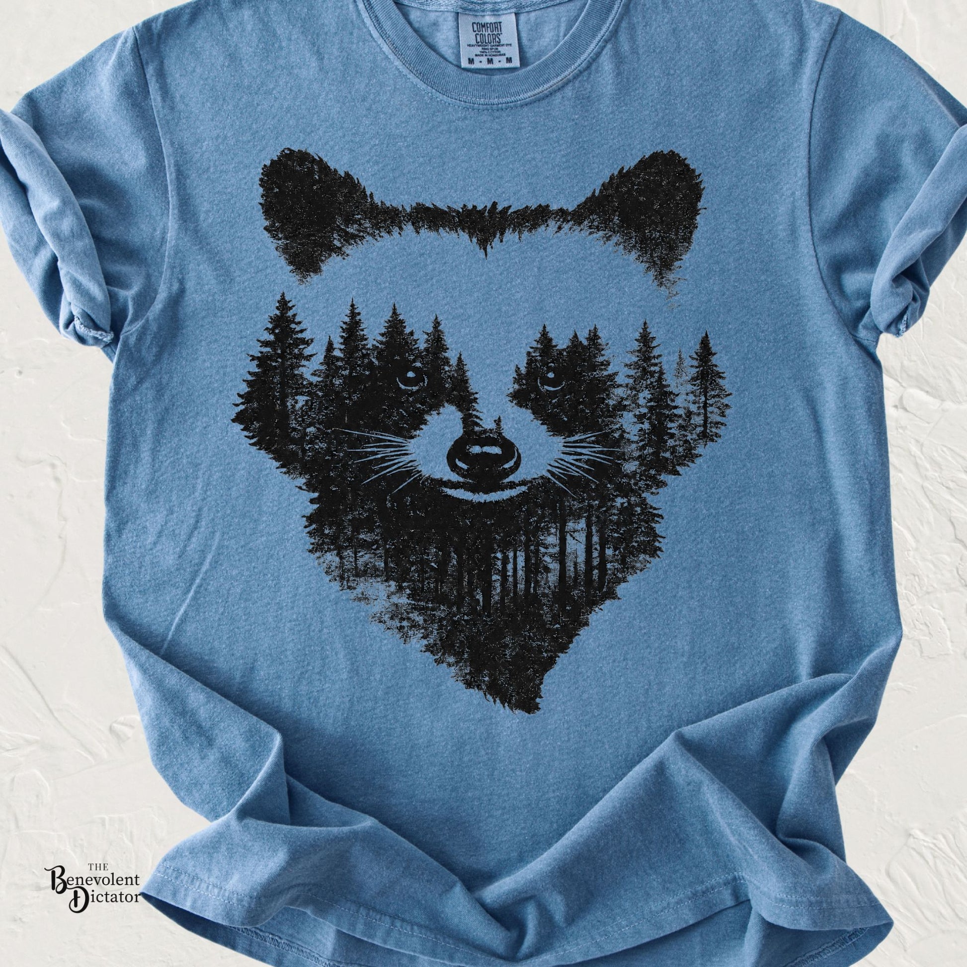 Blue Comfort Colors t-shirt featuring a vintage black silhouette of a raccoon's face, with a pine forest seamlessly forming its facial markings through negative space.