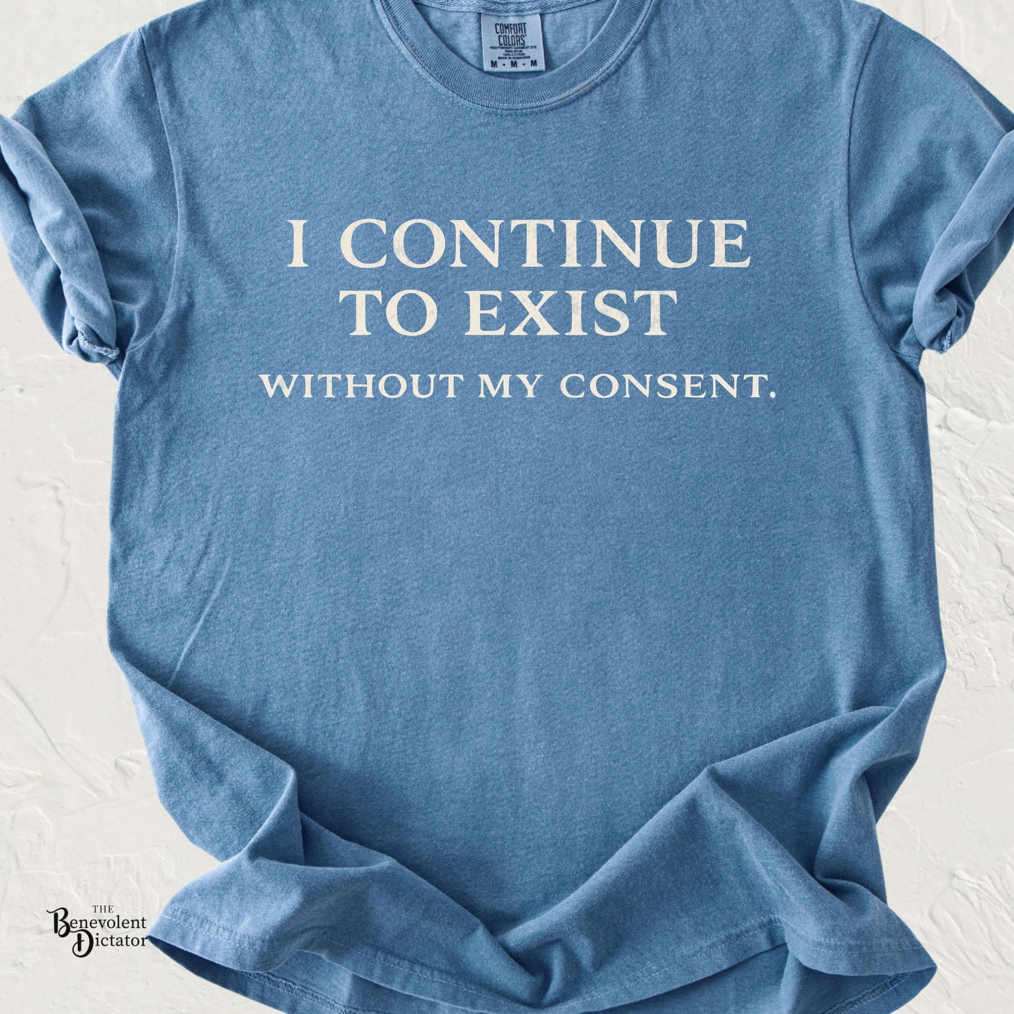 Minimalist typography T-shirt featuring the phrase "I Continue to Exist Without My Consent." printed in a vintage cream serif font on a blue Comfort Colors shirt.