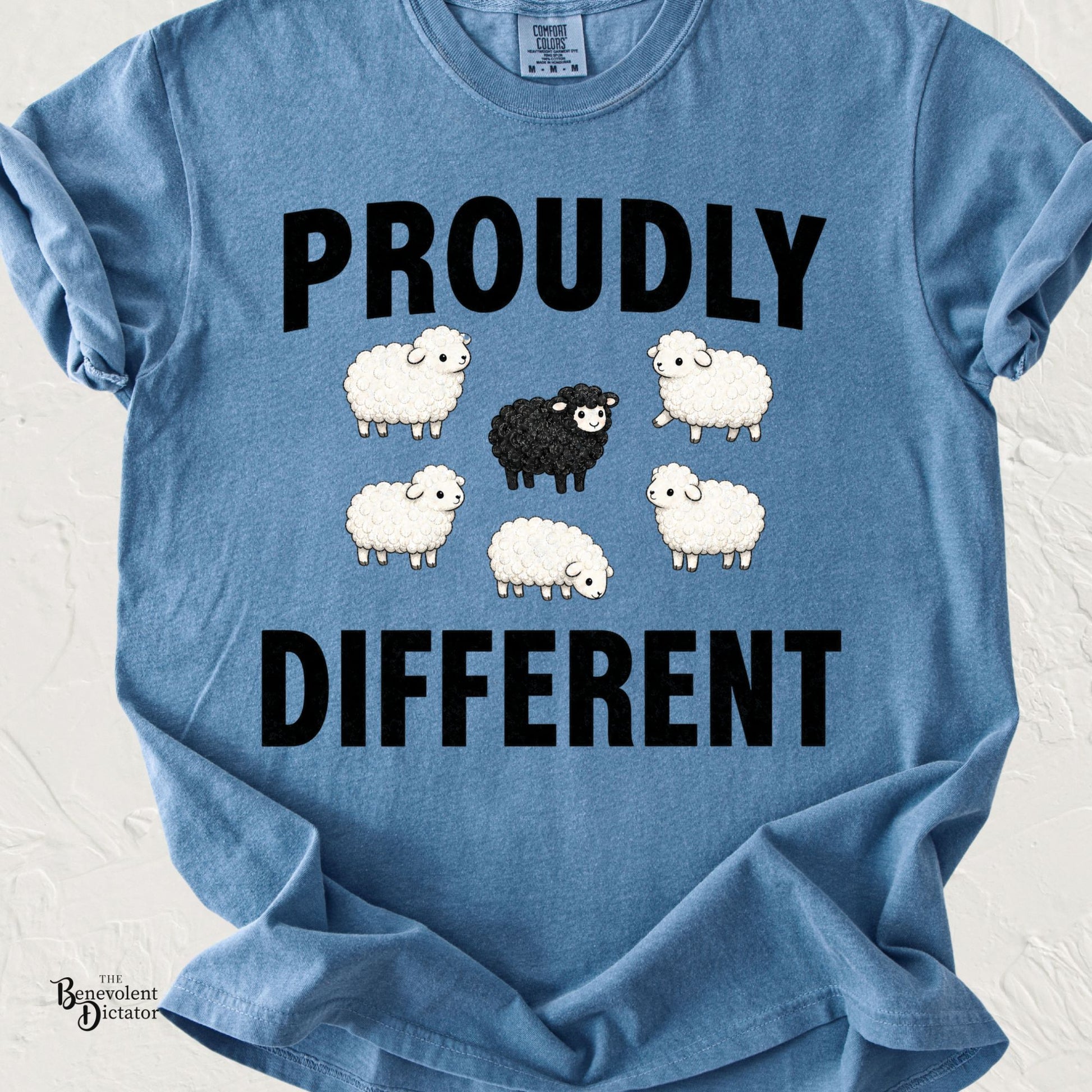 Cute illustration of five white sheep surrounding one smiling black sheep beneath the words "Proudly Different" on a Comfort Colors® t-shirt.