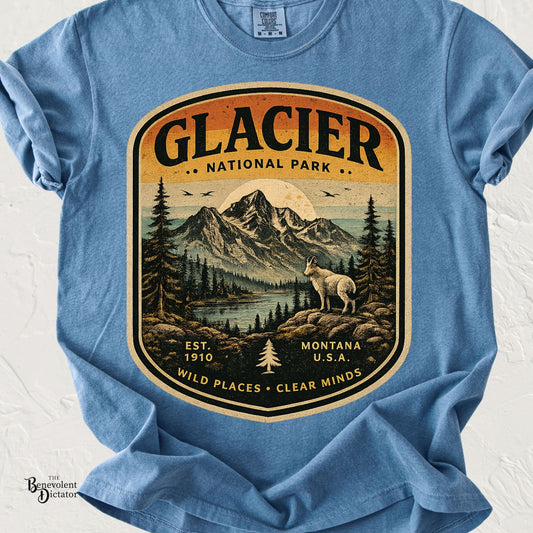 A light blue crewneck t-shirt featuring a large vintage-style graphic print of Glacier National Park with a mountain, lake, pine trees, and a wolf, displayed on a white textured background.