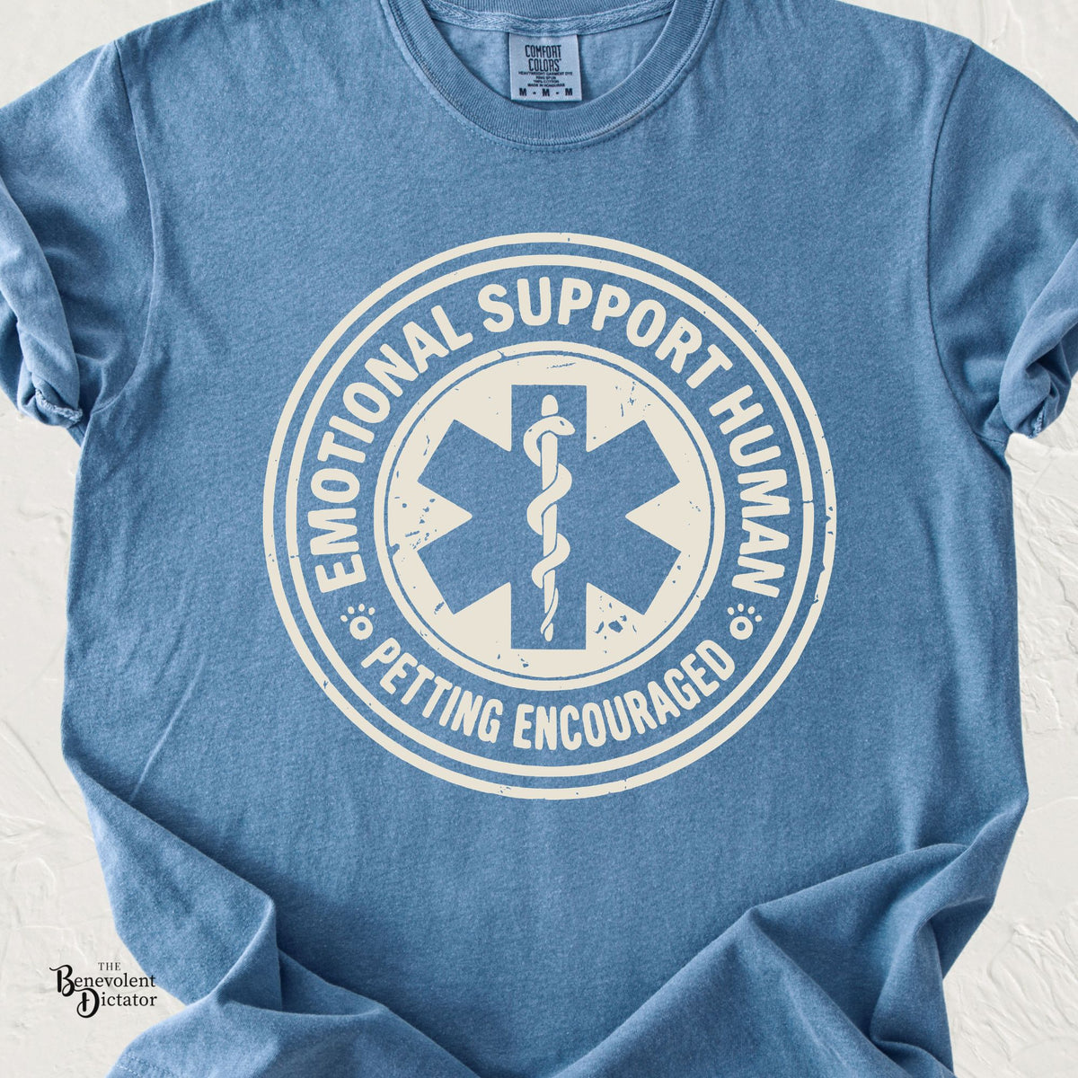Blue Jean Comfort Colors t-shirt featuring a distressed cream circular emblem with a medical Star of Life, a snake-entwined staff, paw prints and the words “Emotional Support Human – Petting Encouraged.”