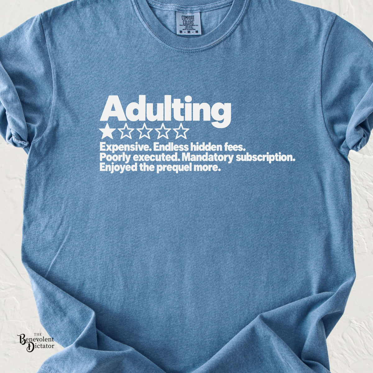 Blue Jean Comfort Colors t-shirt featuring a bold white Adulting one-star review graphic with the text “Expensive. Endless hidden fees. Poorly executed. Mandatory subscription. Enjoyed the prequel more.
