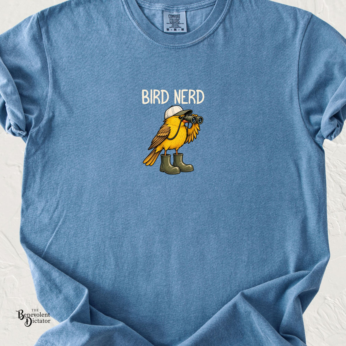 Blue Jean Comfort Colors t-shirt featuring a yellow bird wearing oversized green rain boots and a cap while looking through binoculars with the words “Bird Nerd.”